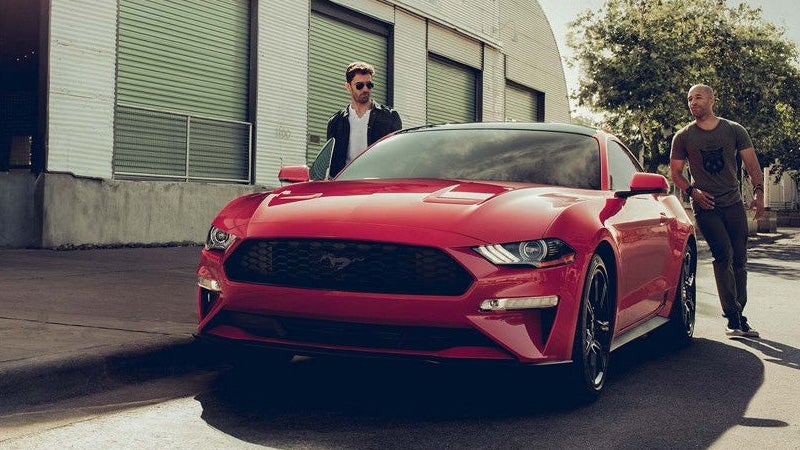 A man stands near the open door of a red Mustang, while another man stands casually on the right.