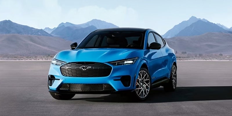A bright blue 2024 Ford Mustang Mach-E electric SUV on an asphalt road with mountains in the background.