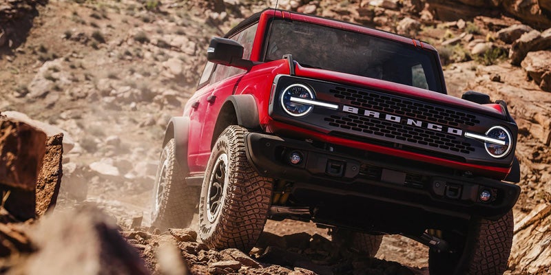 A red Ford Bronco driving uphill on a dusty, rocky desert trail.