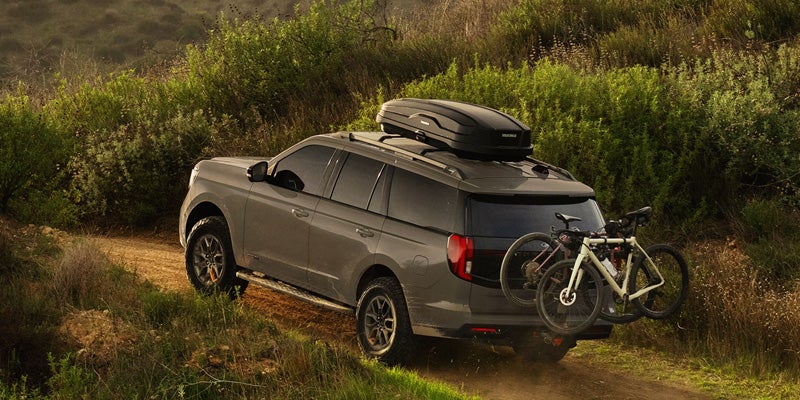 A dark gray SUV with a roof cargo box and two bikes on a rear rack drives up a dirt path surrounded by green brush.
