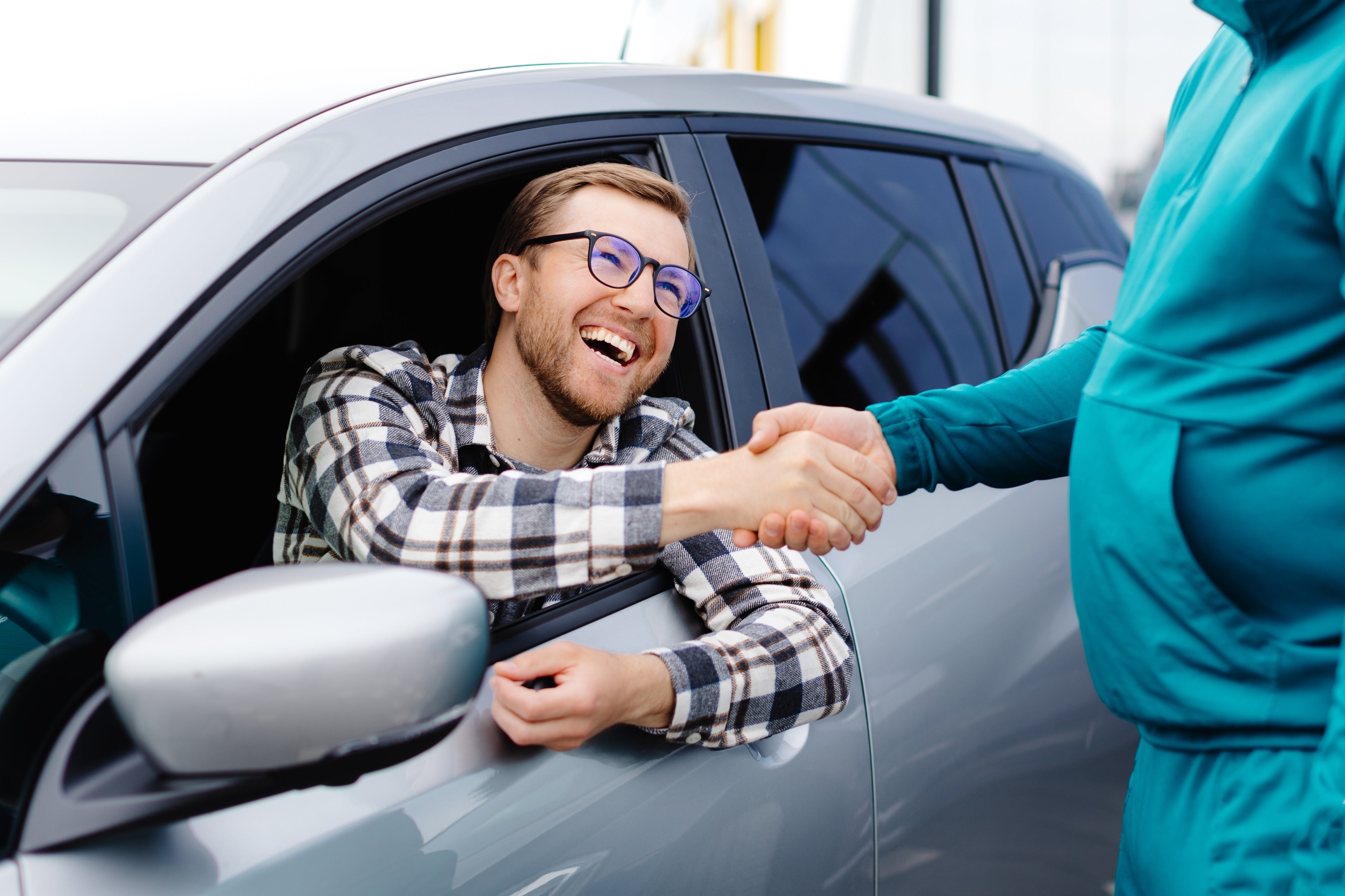 A happy man in a car, wearing glasses and a plaid shirt, shakes hands with another person.