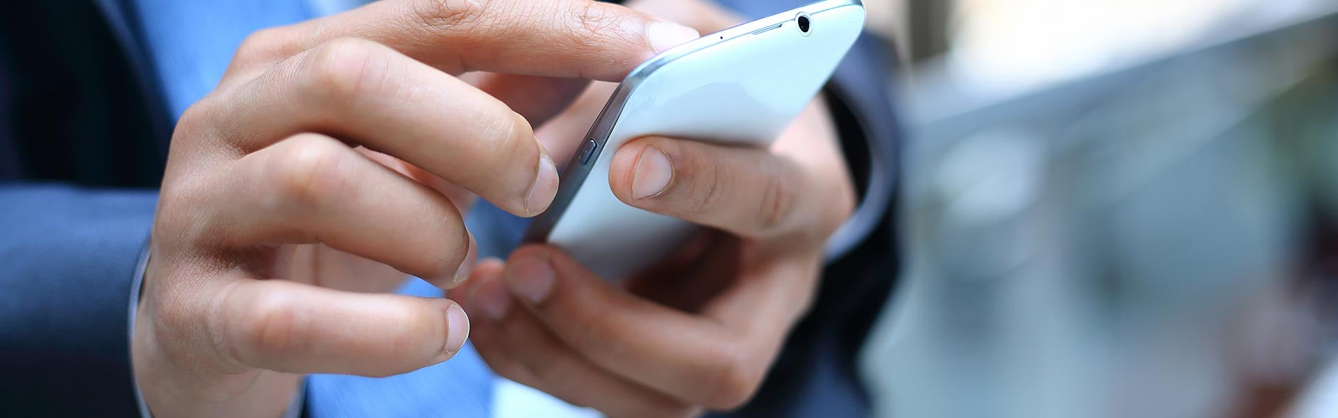 A close-up of hands typing on a white smartphone. The person wears a dark suit with a blue shirt.