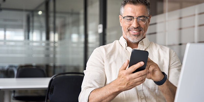 A smiling man with glasses and a beard looks at his phone while sitting at a desk.