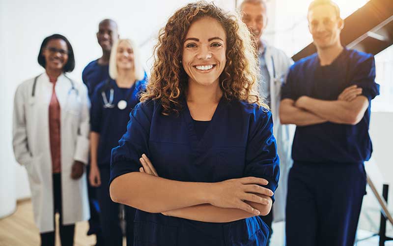 A group of five smiling healthcare professionals, diverse in gender and ethnicity, stand together. A woman in navy scrubs is in front.