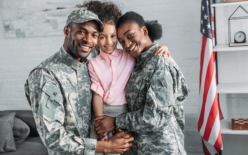 A smiling family, two adults in military camouflage embrace a child in a pink shirt. An American flag is in the background.